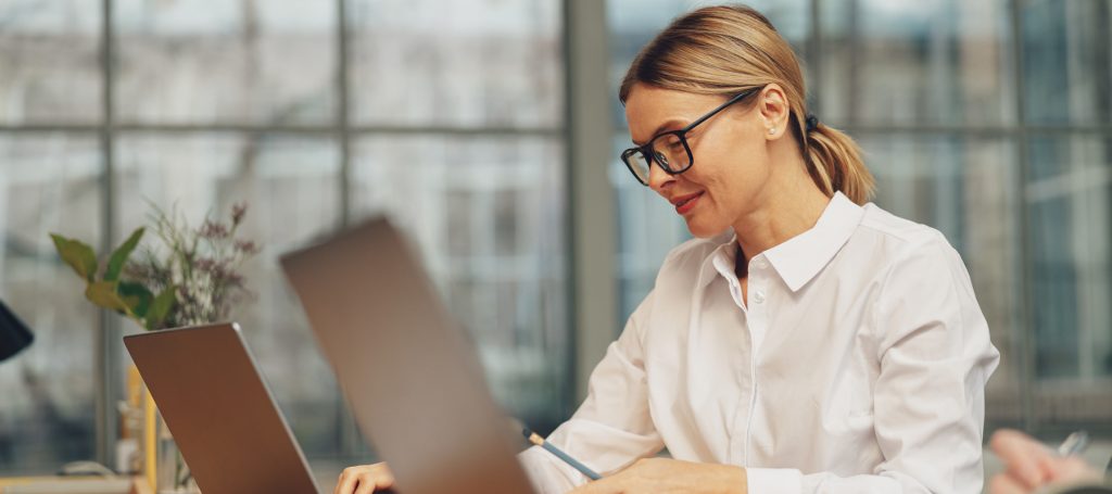 Professional woman wearing glasses and a white shirt, typing on a laptop in a bright office setting.