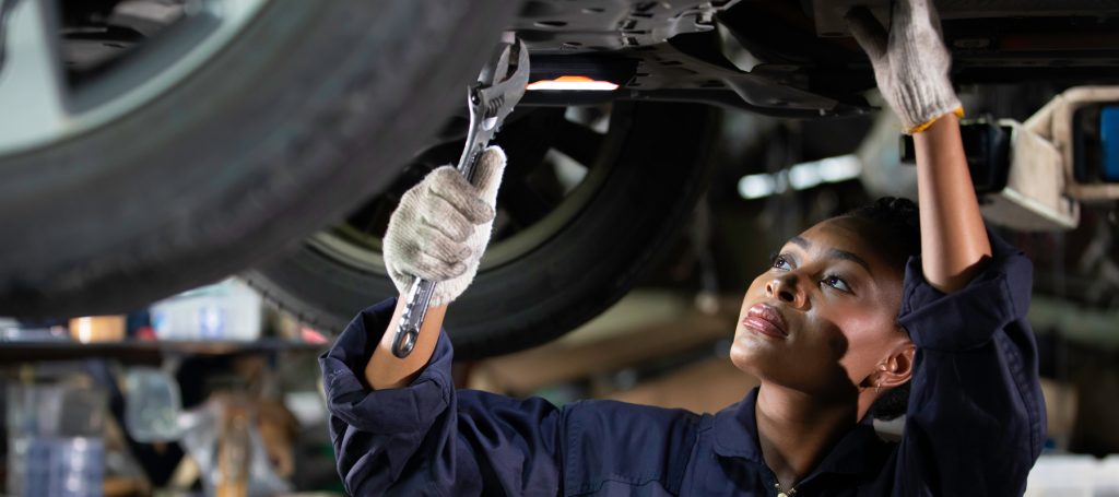 Mechanic wearing gloves and navy coveralls using a wrench under a car lifted on a hoist.